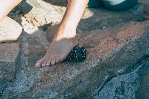 A barefoot person standing on a rocky surface and rolling their foot with a small fascia roller.