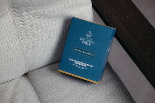 Auf einem beigen Kissen steht eine blaue Schachtel mit der Aufschrift „Vitamin D Essential Wellbeing Check“.