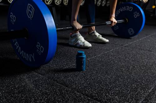 O persoană care se pregătește la sala de fitness să ridice o bară cu discuri albastre; lângă ea, pe podea, se află o sticlă cu suplimente alimentare.