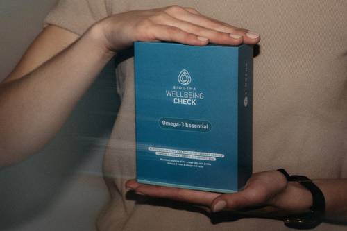 Hände, die eine blaue Schachtel mit der Aufschrift „Wellbeing Check Omega-3 Essential“ und einem tropfenförmigen Logo auf neutralem Hintergrund halten.
