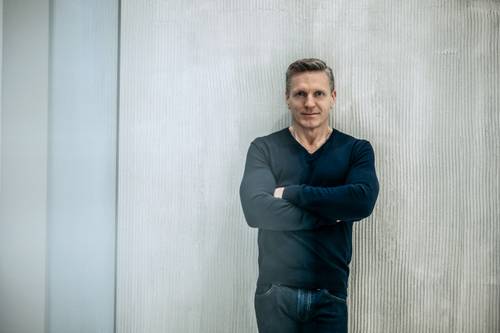 Ein Mann in schwarzem Pullover und Jeans steht mit verschränkten Armen vor einer hell gestrichenen Wand und blickt selbstbewusst in die Kamera.