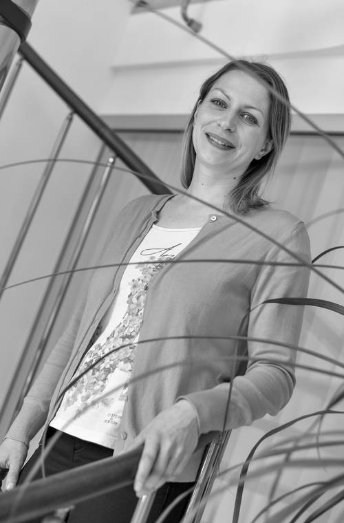 Frau lächelt, steht auf einer Treppe, trägt eine Strickjacke und ein bedrucktes Hemd. Schwarz-Weiß-Bild mit Pflanzenblättern im Vordergrund.