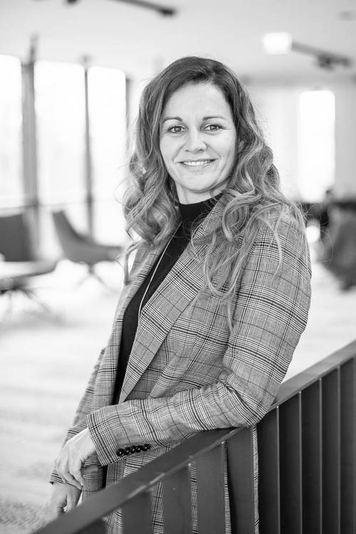 Lächelnde Frau mit welligem Haar in einem gemusterten Blazer, die sich in einem hell erleuchteten Raum an ein Geländer lehnt. Schwarz-Weiß-Foto.