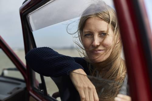 Eine Frau mit langen Haaren sitzt in einer offenen Autotür und lächelt leicht, während der Wind ihr durch die Haare weht; im Hintergrund ist ein Strand zu sehen.