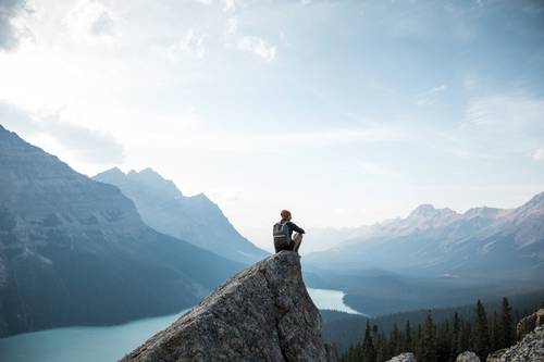 Eine Person sitzt auf einem Felsen und blickt auf eine weite Berglandschaft mit einem See und einem klaren Himmel im Hintergrund.