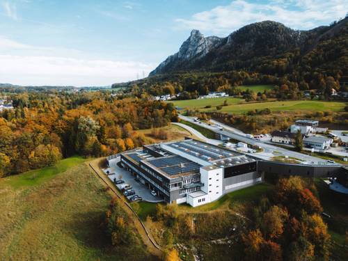 Luftaufnahme des Gebäudes BIOGENA Good Health World, umgeben von Herbstbäumen, einer kurvigen Straße und entfernten Bergen unter einem partly cloudy Himmel.