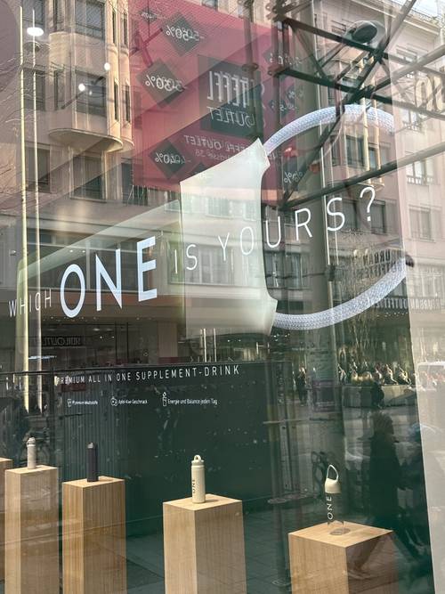 Schaufensterauslage mit verschiedenen Flaschen auf Sockeln, mit dem Text „Which ONE is yours?“ auf dem Glas.