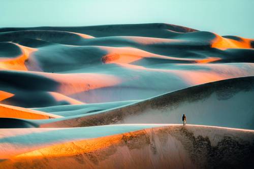 Eine einsame Gestalt wandert über Sanddünen, die von den dramatischen goldenen und blauen Farbtönen der untergehenden Sonne beleuchtet werden, unter einem klaren Himmel.