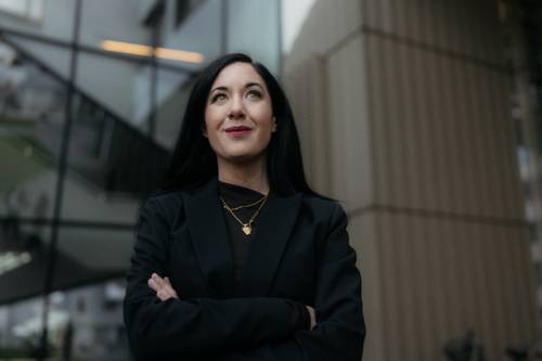 Eine selbstbewusste Frau mit langen dunklen Haaren in einem schwarzen Blazer steht mit verschränkten Armen vor einem modernen Glasgebäude und blickt nach oben.