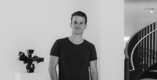 Eine Person in einem schwarzen T-Shirt steht in einem minimalistischen Raum mit einer modernen Skulptur auf einem Sockel und einer geschwungenen Treppe im Hintergrund.