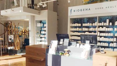 Ein modernes BIOGENA-Geschäftsinterieur mit Holzakzenten, Regale gefüllt mit weißen Nahrungsergänzungsmittel-Flaschen und ein Tresen mit zwei Computerbildschirmen.