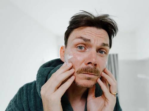 Mann in einem grünen Bademantel trägt Creme auf sein Gesicht in einem hellen Badezimmer auf und schaut in den Spiegel.