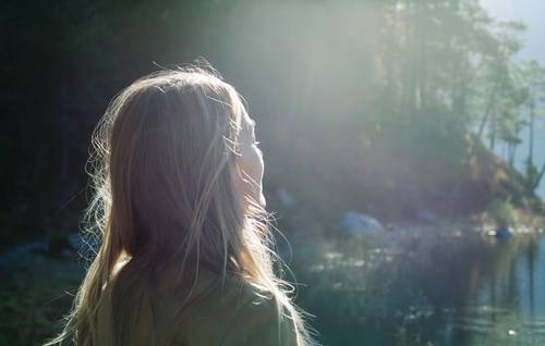 A woman with long hair stands by a calm lake, gazing at the sunlight streaming through the trees.