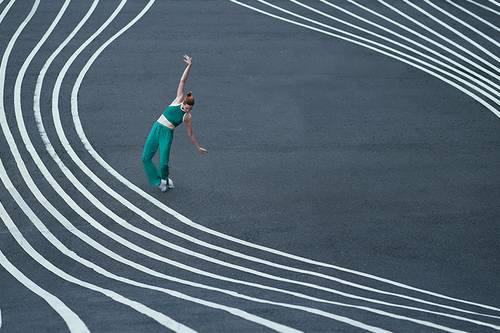 O persoană îmbrăcată în verde dansează pe o stradă cu linii albe ondulate, creând astfel o scenă dinamică și artistică.