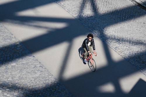 A person is riding a bicycle on a sunlit cobblestone path, with large geometric shadows cast on the ground.