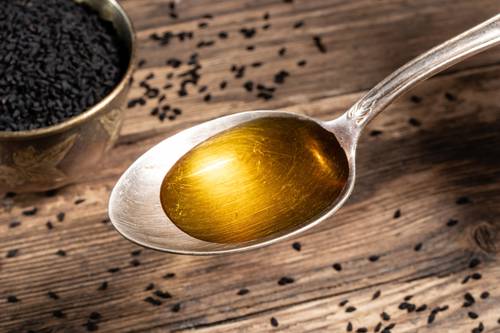 A spoon of golden black cumin oil on a wooden surface, surrounded by scattered black seeds and a bowl of seeds in the background.