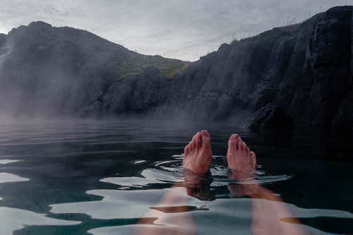 I piedi di una persona immersi in una sorgente termale, circondati da nebbia e colline rocciose sotto un cielo nuvoloso.