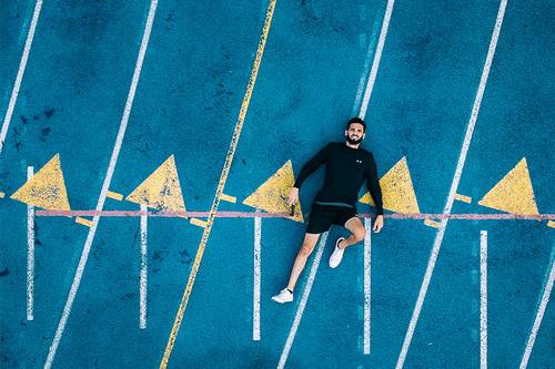 Mann in Sportkleidung liegt auf einer blauen Laufbahn mit gelben Dreieck-Markierungen und weißen Linien, lächelt und wirkt entspannt.