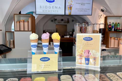 Drei Eistüten in verschiedenen Geschmacksrichtungen stehen in einer Ladentheke, im Hintergrund sind eine Speisekarte und verschiedene Toppings zu sehen.