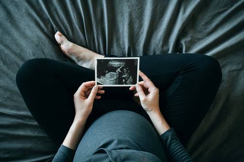 Schwangere Person sitzt mit gekreuzten Beinen auf einer grauen Bettdecke und hält ein Ultraschallbild über ihren Bauch.