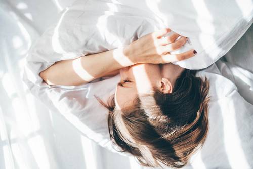Une personne est allongée dans un lit sous des draps blancs, la lumière du soleil et les ombres dessinent des motifs sur son visage et ses cheveux.