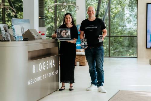Zwei Personen stehen in einem modernen Büro; die eine hält einen Laptop in der Hand, auf dem ein Videoanruf zu sehen ist, die andere hält mehrere verpackte Produkte in der Hand, auf deren Verpackung „BIOGENA“ steht.
