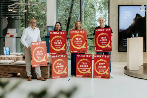 Vier Leute stehen drinnen und halten große rote Plakate hoch, auf denen „TEST SIEGER 2026“ steht – ein Zeichen für eine Leistung oder eine Auszeichnung.