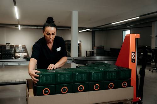 A woman in a warehouse is sorting green crates labelled ‘ONE’ using a pallet truck.