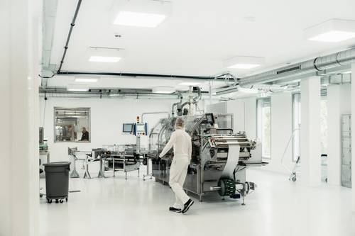 A person wearing white protective clothing operates machinery in a clean, white industrial laboratory with pipework running along the ceiling and bright lighting.