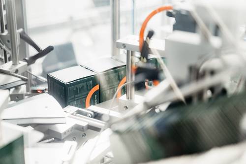 A close-up of a packaging machine in a factory, processing dark green boxes with orange details.