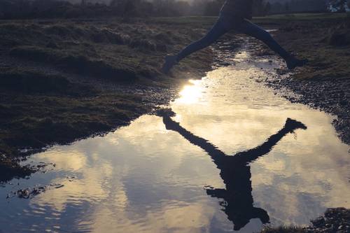 A person jumps over a reflective puddle at sunset, creating a mirrored silhouette against the sky.