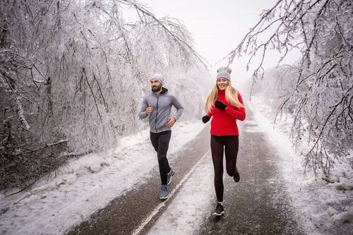 Two people are jogging on a snow-covered path surrounded by frost-covered trees, dressed in winter sports clothing.