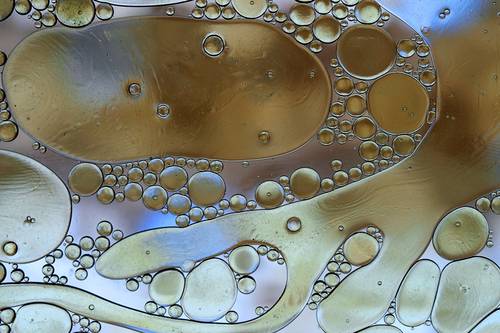 Close-up of oil droplets in water forming bubbles of varying sizes with a shimmering, abstract pattern against a blue-brown background.
