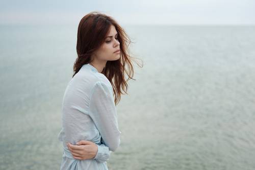 Una donna dai lunghi capelli è in piedi in riva al mare, indossa un vestito azzurro e sembra pensierosa mentre una leggera brezza soffia.