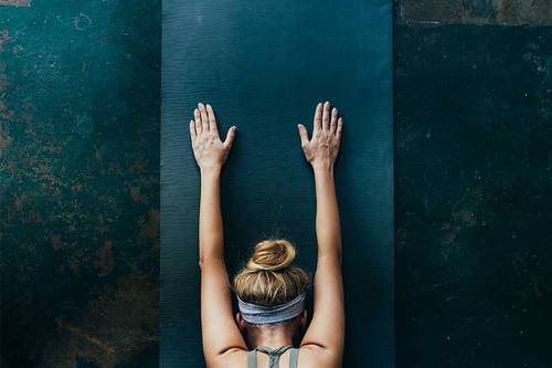 Mujer en una postura de yoga sobre una esterilla, con los brazos extendidos hacia delante, el pelo recogido en un moño y una cinta en la frente, vista desde arriba.