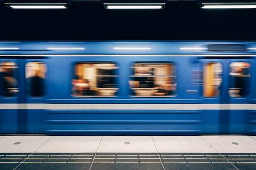 Un metrou albastru trece cu viteză printr-o stație subterană, în care se văd pasageri neclari.