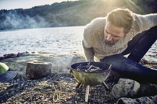 A person wearing a jumper lights a small fire in a portable barbecue on the lakeshore, surrounded by rocks and nature, with sunlight in the background.