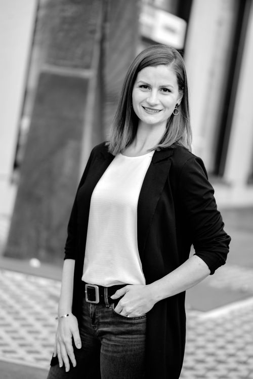 Eine Frau in einem schwarzen Blazer und einem weißen Oberteil steht lächelnd im Freien. Der Hintergrund ist unscharf, wodurch ihre selbstbewusste Haltung hervorgehoben wird. Schwarz-Weiß-Foto.