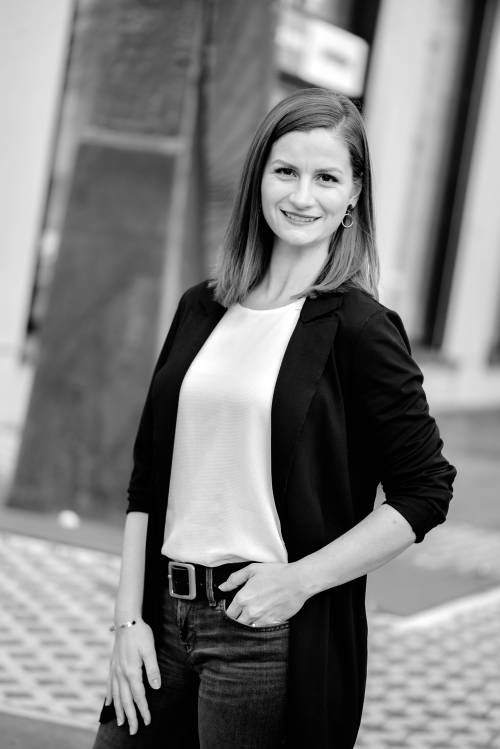 Eine Frau in einem schwarzen Blazer und einem weißen Oberteil steht lächelnd im Freien. Der Hintergrund ist unscharf, wodurch ihre selbstbewusste Haltung hervorgehoben wird. Schwarz-Weiß-Foto.