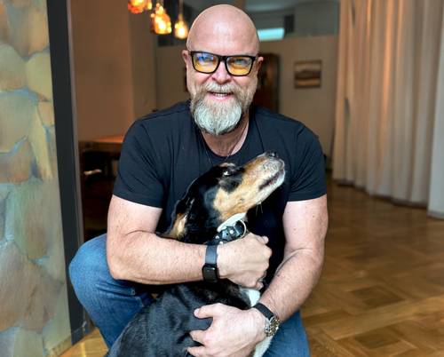 Mann mit Brille und Bart lächelt, während er einen schwarz-braunen Hund in einem Raum umarmt.