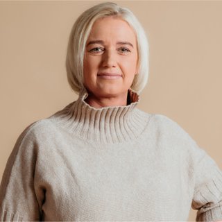 Frau mit kurzen blonden Haaren, die einen beigen Rollkragenpullover trägt, vor einem hellbraunen Hintergrund steht und sanft lächelt.