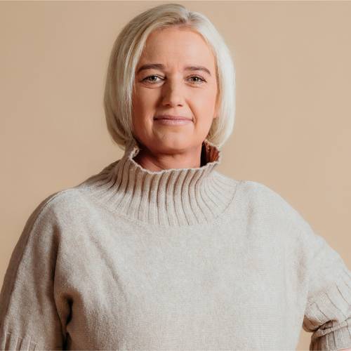 Frau mit kurzen blonden Haaren, die einen beigen Rollkragenpullover trägt, vor einem hellbraunen Hintergrund steht und sanft lächelt.