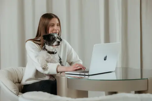 Eine Frau in einem weißen Pullover lächelt, während sie an einem Glastisch sitzt und einen Laptop benutzt. Auf ihrem Schoß sitzt ein kleiner schwarz-weißer Hund.