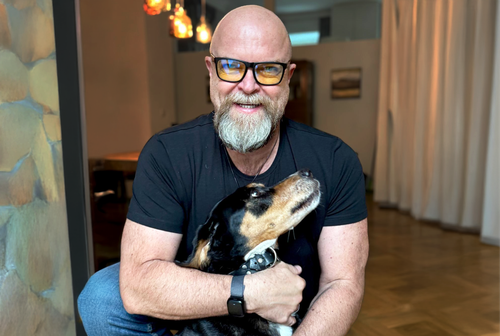 Ein lächelnder Mann mit Glatze und Brille kniet da und umarmt einen schwarz-braunen Hund in einem Raum mit warmer Beleuchtung und unscharfem Hintergrund.