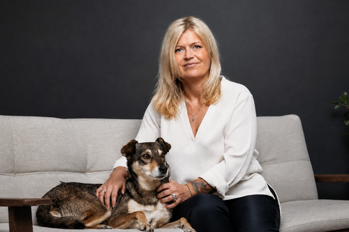 Eine Frau mit blonden Haaren sitzt auf einem Sofa und streichelt einen braun-schwarzen Hund; beide blicken in die Kamera, vor einem dunklen Hintergrund.