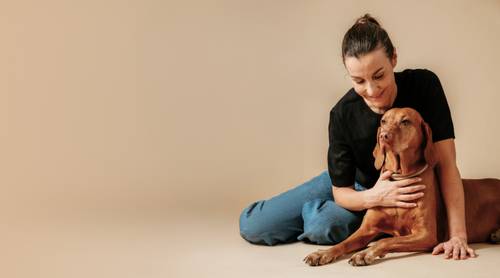Frau sitzt auf dem Boden und umarmt liebevoll einen braunen Hund vor einem beigen Hintergrund.