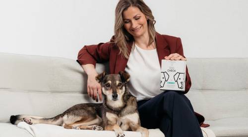 Tanja Fleischanderl von BIOGENA PETS sitzt in einem roten Blazer mit einem Hund auf einem Sofa und hält eine „DNA Dog Check" Box, lächelnd.