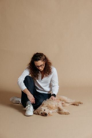 Eine Frau mit lockigem Haar und Brille sitzt auf dem Boden und streichelt einen flauschigen Hund, der auf dem Rücken liegt