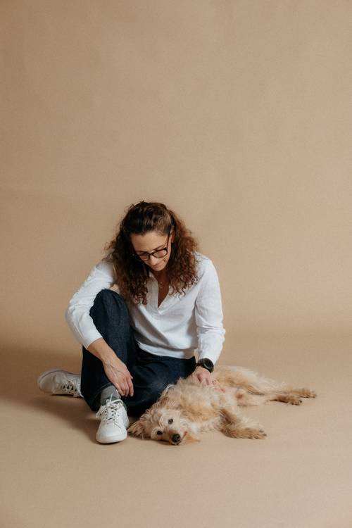 Eine Frau mit lockigem Haar und Brille sitzt auf dem Boden und streichelt einen flauschigen Hund, der auf dem Rücken liegt