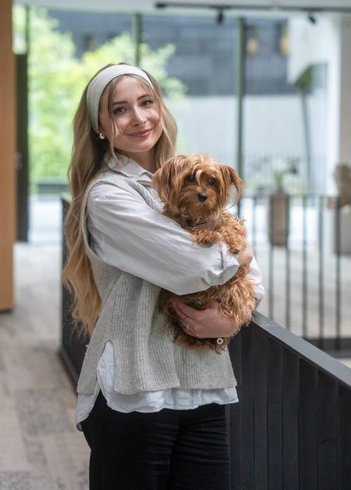 Eine Frau mit langen Haaren und einem Stirnband hält einen kleinen, flauschigen Hund in einem Raum und lächelt sanft in die Kamera